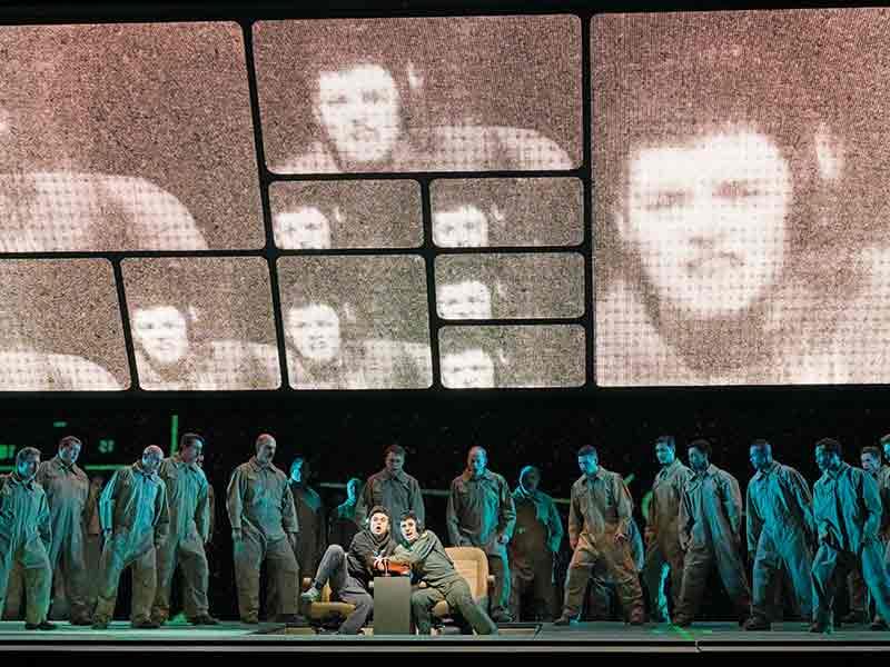 The Met Opera production of Grounded, showing the singers with large digital closeups on screens behind them